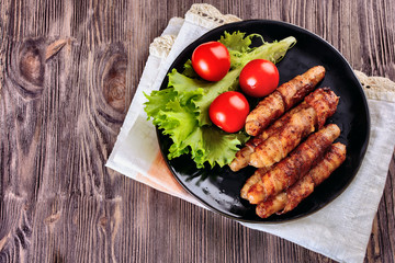 Appetizing sausages wrapped in bacon and grilled next to tomatoes and lettuce are on a wooden table, top view