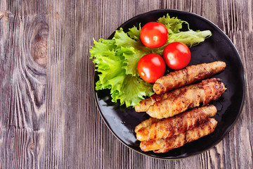 Appetizing sausages wrapped in bacon and grilled next to tomatoes and lettuce are on a wooden table, top view
