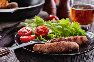 Appetizing sausages wrapped in bacon and grilled next to tomatoes, lettuce and and glass of beer are on a wooden table