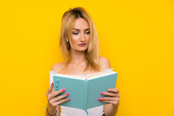 Young blonde woman over isolated yellow wall holding and reading a book