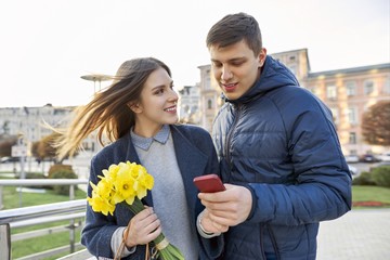 Outdoor portrait of beautiful romantic couple, young man and woman with bouquet of yellow flowers of daffodils and looking in smartphone, spring city background