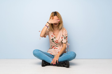 Young blonde woman sitting on the floor covering eyes by hands. Do not want to see something
