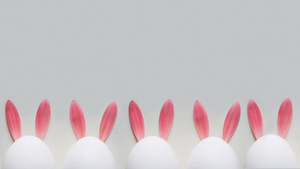 Four Easter white eggs with rabbit ears made of pastel pink gerbera petals on grey background