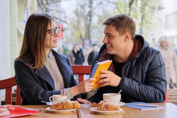 Young couple of students study in outdoor cafe, drink coffee tea, eat croissants, background is spring city street