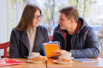 Young couple of students study in outdoor cafe, drink coffee tea, eat croissants, background is spring city street