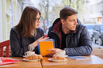 Fototapeta premium Young couple of students study in outdoor cafe, drink coffee tea, eat croissants, background is spring city street