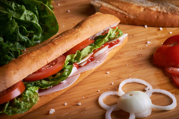 Fresh baguette sandwich with ham, tomato, onion, lettuce and ketchup surrounded by some ingredient on a bamboo breadboard
