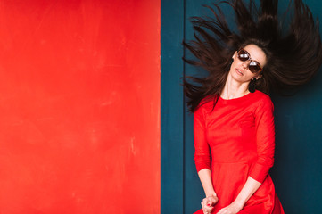 Beautiful fashion girl with long hair, spanish appearance in sunglasses and red elegnat dress posing on blue red wall in studio. Stylish brunette babe with flying hair. Professional hairstyle concept.