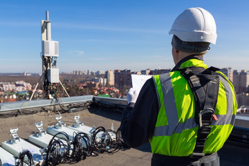 Professional industrial climber in helmet and uniform reads technical documentation and antennas of GSM DCS UMTS LTE bands, outdoor radio units on the roof. Working process of upgrading