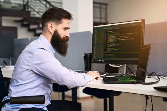 Software programming. Bearded man working on computer in IT office, sitting at desk writing code, working on a project in software development company or startup. High quality image.