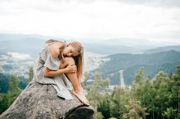 Naklejka na ściany i meble Lonely young girl sits at stone on top of mountain with far view at hills and cloudy sky and hugs her legs with head lying on knees. Beautiful blonde woman outdoor summer portrait. Female at nature.