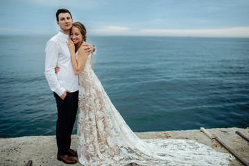 Beautiful lovers newlyweds posing near the sea at sunset. The bride in a luxurious dress, the groom in a stylish suit