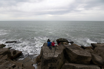 People sit on a rock by the sea on a cloudy day, Mar del Plata, Buenos Aires, Argentina.