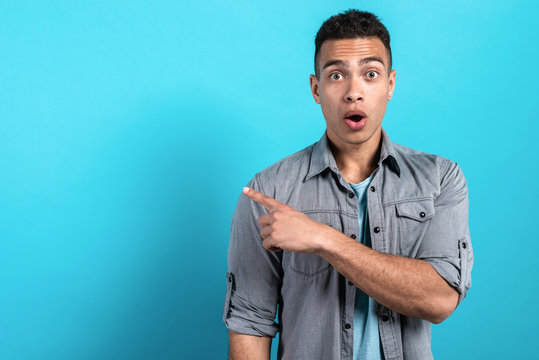 Young surprised man pointing his hands sideways and standing against a blue studio background. - Image
