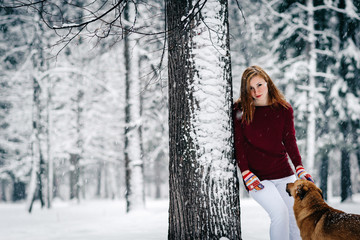 a girl in a Burgundy sweater and white pants stands leaning against a tree near the Red Dog amid the snowcovered forest