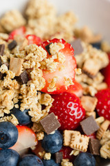 Healthy muesli or cereal breakfast bowl with granola made of toasted rolled oats, organic honey, milk chocolate chips and tiny graham cracker cookies served with fresh strawberries and blueberries