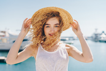 Beautiful young curly woman blonde on the background of the sea and the water holds on to the edges of the hat and laughs