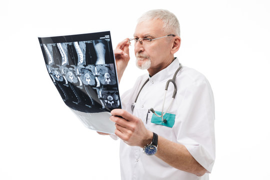 Old thoughtful doctor with gray hair and beard in eyeglasses and uniform with stethoscope on neck looking at X-ray image while spending time over white background