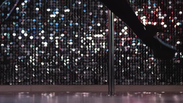 Young stripper in black dances sexually with pylone in night men's club. Poledance. Close-up legs of unrecognizable woman doing tricks with pole.