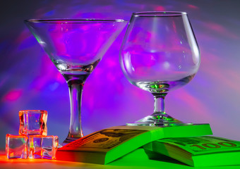 Martini Glass together with Cognac Glass on a packs of the 100s of us dollars and ice cubes with flashing bright violet lights on background