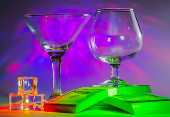 Martini Glass together with Cognac Glass on a packs of the 100s of us dollars and ice cubes with flashing bright violet lights on background