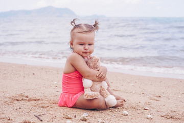 pensive little girl hugging teddy bear and looking away while sitting on seashore.