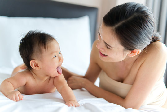 Asian Cute Baby Crawling And Playing With Her Beautiful Mother On The Bed. They Are Looking On Each Other And Mother Is Smiling When Baby Sticking Tongue Out . Touch Of Love From Mother Concept.