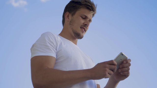 Smiling young guy messaging on smartphone outdoor. Cheerful bearded guy using modern device against blue sky. Technology concept