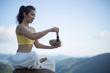 Young woman doing exercise yoga. sitting on the rock and playing on a tibetian singing bowl in the morning, (The Nepal word on the tibetan singing bowls means wish everything is fine.)