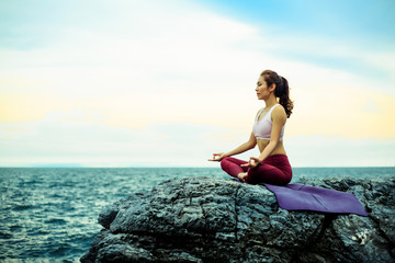 Fototapeta na wymiar Young woman doing exercise yoga. sitting on the rock, doing Padmasana exercise, Lotus pose. Amazing yoga landscape in beautiful sky and enjoying sea view with evening.