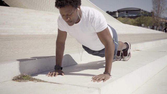 Sporty young man listening music during workout. Confident smiling African American sportsman doing push ups on stairs. Concept of sport