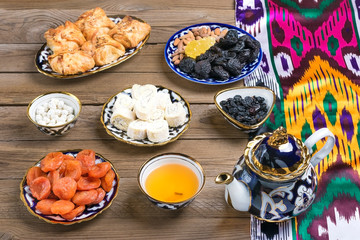 Traditional Uzbek sweets - dried apricot, rohat Turkish delight, raisin, samsa, almond, teapot with tea and bowl with national ornament on wooden table. Flat lay.