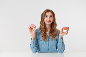 Confused young pretty woman with long blond wavy hair, sitting at the table and holds donuts in her hands. Looks up and wonders whether to eat sweets for the night. isolated over white background.