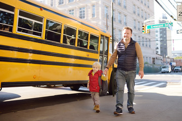 Elementary student hold hands his father near yellow school bus on background.