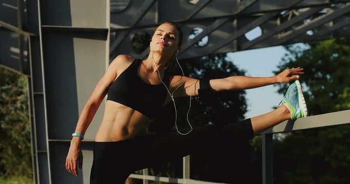 Portrait shot of the charming young Caucasian cheerful woman listening to the music in the headphones and stretching her body, legs and arms on a sunny day.