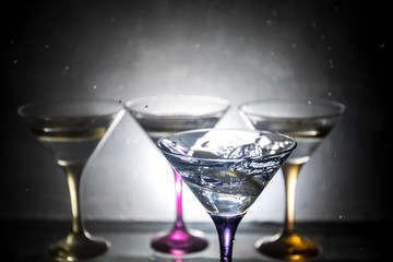 Martini cocktail glass in hand splashing on dark toned smoky background or colorful cocktail in glass with splashes and olives. Party club entertainment. Mixed light. Selective focus
