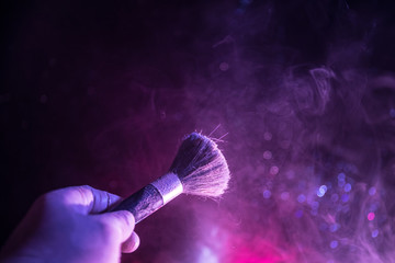 Makeup brush in hand with cosmetic powder on dark background with light and smoke. Powder splash on dark