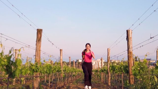 Bellissima ragazza con maglia rosa e pantaloni neri corre allegramente in campagna tra le vigne.