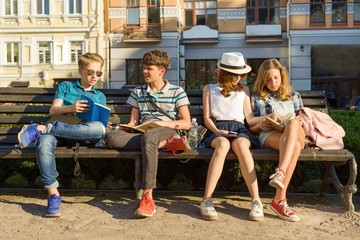 Fototapeta premium Happy 4 teenage friends or high school students are having fun, talking, reading phone, book. Friendship and people concept, city street background