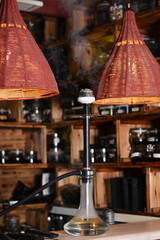 Luxurious long black matte hookah with clear water in bowl with coals on a beige bar counter next to large red lamps made of cloth. The smoke is blown out of the metal kaloud.