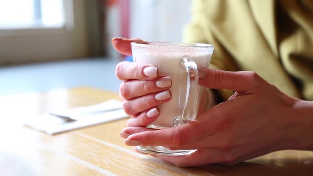A glass beaker with hot foamed milk. Woman warms hands on a glass mug of hot milk in a cafeshop. Beautiful latte with a big foam. A woman touches a glass with a hot latte