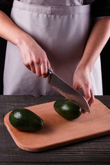 A young woman in an gray apron cuts an avocado on a wooden cutting board