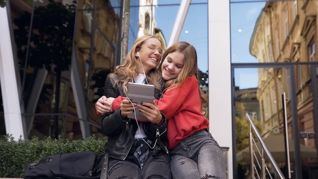 Two women friends using digital tablet in outdoors. Smiling girl friends looking over photos on tablet device, walking with pleasure near the modern center in the city. Outdoors