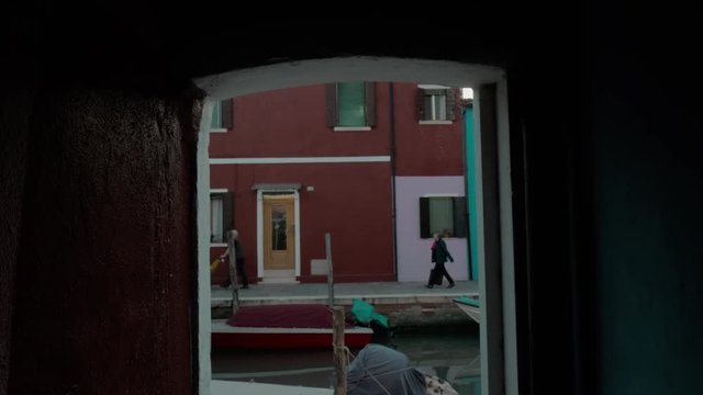 BURANO, ITALY - APRIL 21, 2018: View through the doorway to the canal with boats and some people walking along the houses in the street