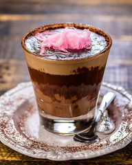 A warm glass of hot chocolate on a white plate with a silver spoon, topped with pink colored fairy floss