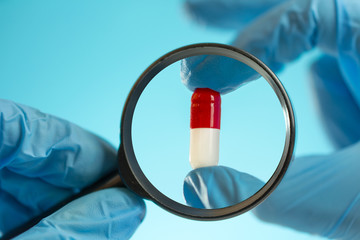 in the hands of a scientist a magnifying glass and a red-white capsule in blue medical gloves on a...