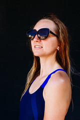 smiling tanned woman in a sports swimsuit and sunglasses stands on a black background. Young woman with wet hair