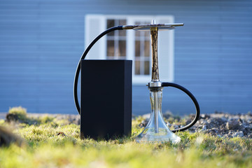 Beautiful beige wooden hookah with lightning patterns from an electric current with a clean bowl with a black box in the sun on the green grass on the background of a blue house with white windows.
