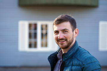 Portrait of a laughing bearded man with freckles with short brown hair and green eyes in a blue jacket and plaid shirt on the background of a blue house with white windows in the sun.