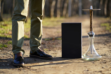 Men's legs in olive trousers and black patent leather shoes with brown laces next to a beige wooden hookah with lightning patterns and a box in the autumn forest at sunset in the rays of the sun.